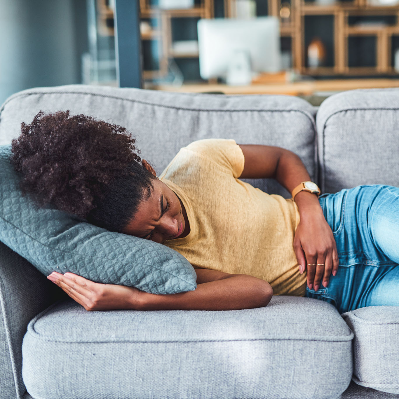 black woman lying down with her hand on her stomach in pain on gray couch